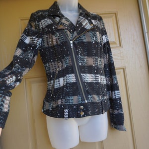 May include: A black and gray patterned jacket with a diagonal zipper closure and a notched collar. The jacket features a belt detail at the waist and a textured fabric with accents of white and teal.