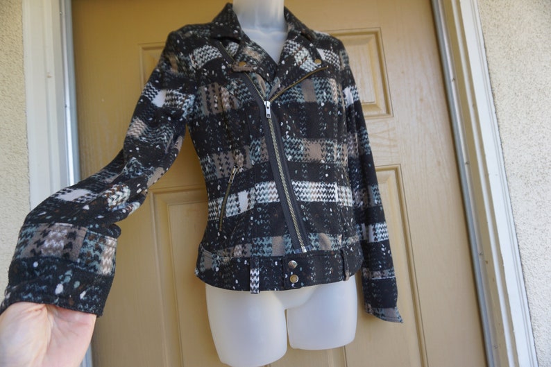 May include: A black and gray patterned jacket with a diagonal zipper closure and a notched collar. The jacket features a belt detail at the waist and a textured fabric with accents of white and teal.