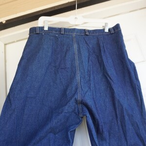 May include: Dark blue denim trousers hanging on a white hanger. The trousers have a high waist, a button and zip closure, and a straight leg design. The denim is a solid, medium-weight fabric.