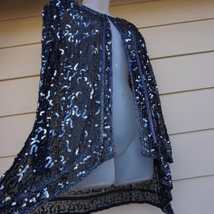 May include: A dark navy blue sequined cape-style jacket with a sheer, flowing design. The jacket is adorned with silver sequins and features a blue trim along the edges. The garment is displayed on a mannequin.