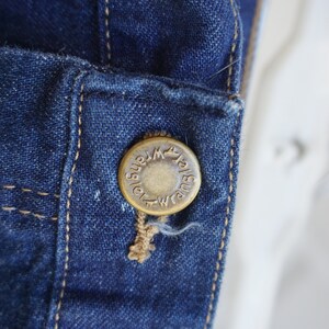 May include: Close-up of a dark blue denim jacket detail. A brass button with the word "Wrangler" is attached to the jacket. The button is sewn onto the denim with tan thread. The denim has visible stitching.