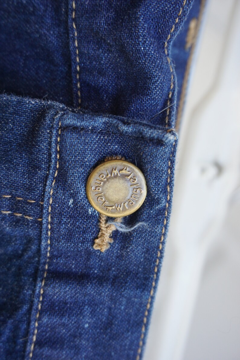 May include: Close-up of a dark blue denim jacket detail. A brass button with the word "Wrangler" is attached to the jacket. The button is sewn onto the denim with tan thread. The denim has visible stitching.