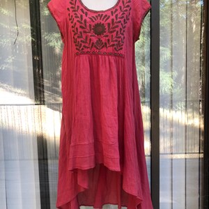 May include: A vibrant pink dress with short sleeves and a flowing, asymmetrical hemline. The dress features intricate brown embroidery around the neckline and on the bodice, showcasing a floral design. The fabric appears lightweight and textured, perfect for warm weather.