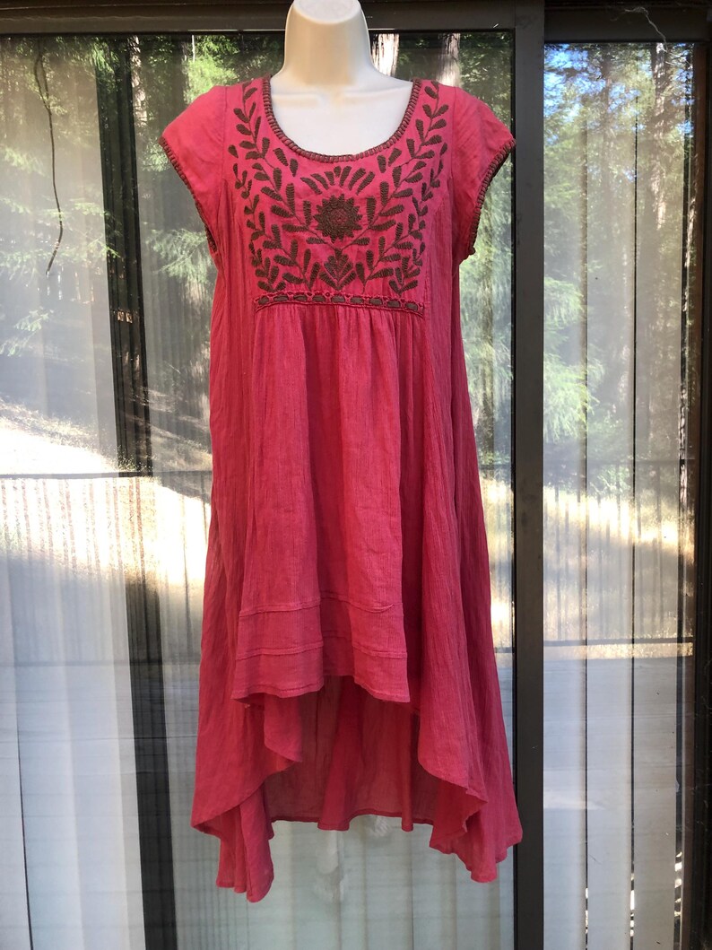 May include: A vibrant pink dress with short sleeves and a flowing, asymmetrical hemline. The dress features intricate brown embroidery around the neckline and on the bodice, showcasing a floral design. The fabric appears lightweight and textured, perfect for warm weather.