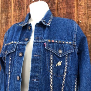 May include: A blue denim jacket with white stitching and colorful embroidery around the collar and pockets.
