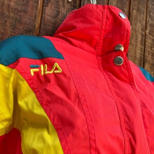 May include: A vintage Fila jacket in red, yellow, and teal. The jacket features the Fila logo embroidered in yellow. The collar has two silver buttons. The jacket is made of a lightweight material, perfect for layering.