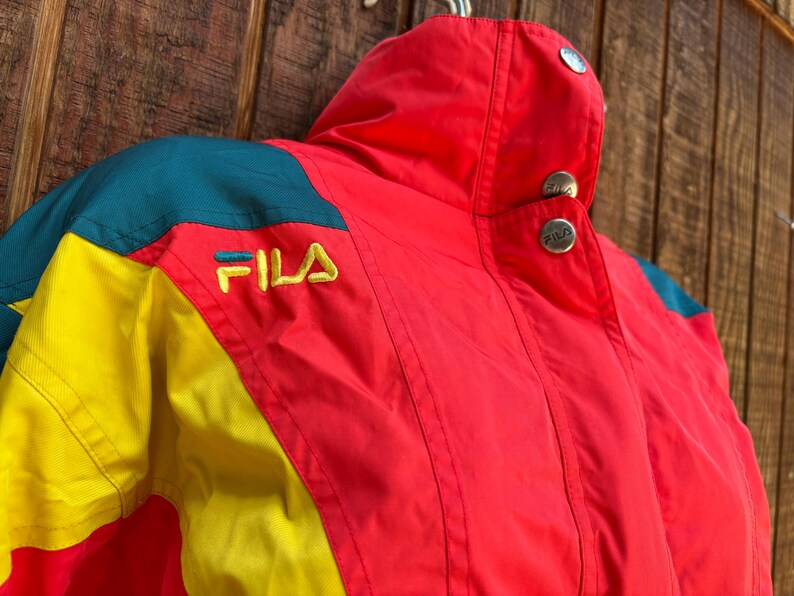 May include: A vintage Fila jacket in red, yellow, and teal. The jacket features the Fila logo embroidered in yellow. The collar has two silver buttons. The jacket is made of a lightweight material, perfect for layering.