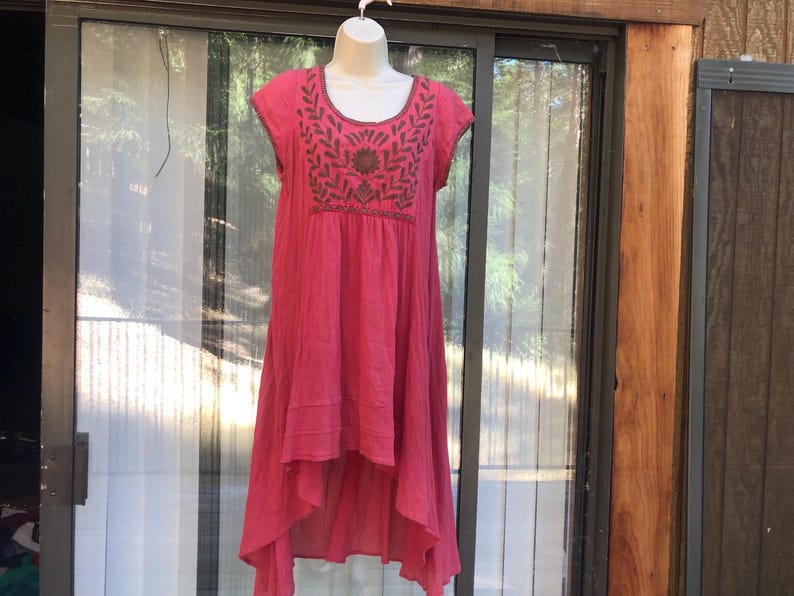 May include: A vibrant pink dress with a high-low hemline and cap sleeves. The dress features intricate embroidery in shades of brown around the neckline and bodice. The dress is displayed on a mannequin.