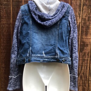 May include: A blue denim jacket with a gray knit hood and sleeves. The jacket has a distressed look with faded areas and a button-up front. The sleeves have a textured pattern and the hood is a solid gray color.