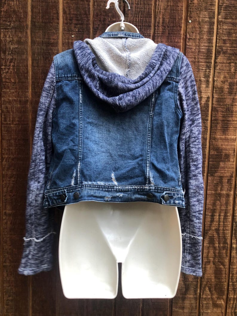 May include: A blue denim jacket with a gray knit hood and sleeves. The jacket has a distressed look with faded areas and a button-up front. The sleeves have a textured pattern and the hood is a solid gray color.