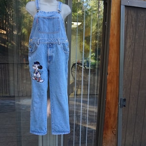 May include: Light blue denim overalls with adjustable straps and a front bib pocket. The left leg features an embroidered Mickey Mouse design. The overalls have a classic, relaxed fit and are displayed on a mannequin.
