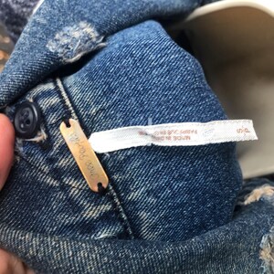 May include: Close-up of a blue denim garment with distressed details. A small, rectangular copper-colored tag with the brand name "Free People" is attached. A white tag with text is also visible. The garment has a button closure.