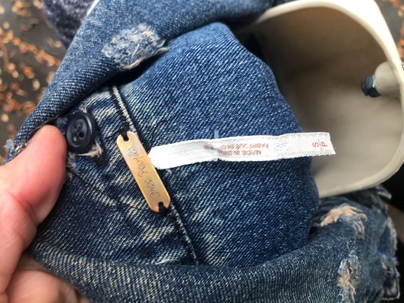 May include: Close-up of a blue denim garment with distressed details. A small, rectangular copper-colored tag with the brand name "Free People" is attached. A white tag with text is also visible. The garment has a button closure.