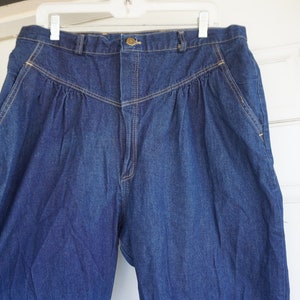 May include: Dark blue denim trousers with a high waist and pleated front. The trousers have a button and zip closure, with pockets on the sides and back. The stitching is a contrasting tan colour. The trousers are hanging on a white hanger.