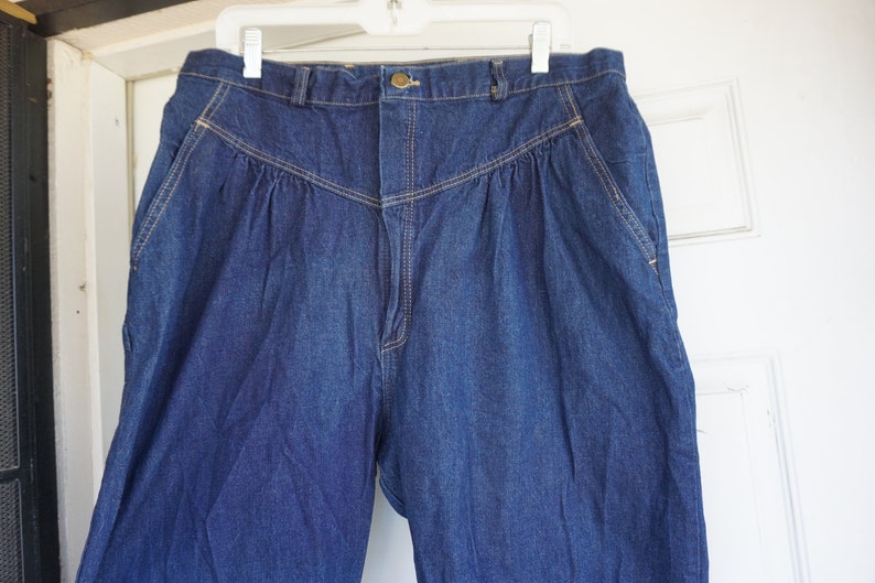 May include: Dark blue denim trousers with a high waist and pleated front. The trousers have a button and zip closure, with pockets on the sides and back. The stitching is a contrasting tan colour. The trousers are hanging on a white hanger.