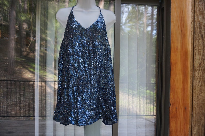 K&ouml;nnte beinhalten: Ein marineblaues Paillettenkleid mit Neckholder-Ausschnitt und V-Ausschnitt. Das Kleid ist &auml;rmellos und locker fallend. Die Pailletten verleihen dem Kleid ein schimmerndes Aussehen. Das Kleid wird auf einer wei&szlig;en Schaufensterpuppe pr&auml;sentiert.