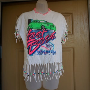 May include: White t-shirt with a graphic of a green car and the words "Fast Bash" in pink and blue. The shirt has fringe detailing and colorful beads. The text "Reno, Nevada, Labor Day 1991" is also visible.