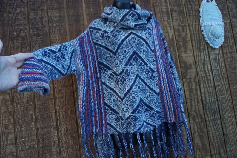 May include: A blue, white, and red patterned poncho with a cowl neck and fringe. The poncho features a geometric design and is made of a woven fabric. The sleeves have a striped pattern in red, white, and blue.