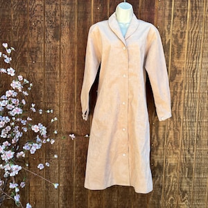 May include: A long-sleeved, knee-length coat in a solid light peach color. The coat features a notched lapel collar, a button-down front closure with five visible buttons, and straight sleeves with buttoned cuffs, displayed on a mannequin.