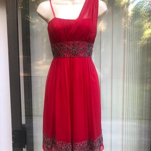 K&ouml;nnte beinhalten: Ein leuchtend rotes One-Shoulder-Kleid mit einem bestickten Bund und Saum. Das Kleid hat einen flie&szlig;enden, knielangen Rock und einen zarten Tr&auml;ger auf einer Schulter. Das Oberteil ist figurbetont, und das Kleid wird auf einer Schaufensterpuppe pr&auml;sentiert.