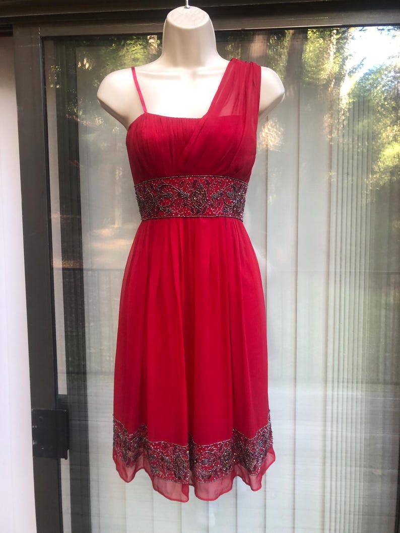 K&ouml;nnte beinhalten: Ein leuchtend rotes One-Shoulder-Kleid mit einem bestickten Bund und Saum. Das Kleid hat einen flie&szlig;enden, knielangen Rock und einen zarten Tr&auml;ger auf einer Schulter. Das Oberteil ist figurbetont, und das Kleid wird auf einer Schaufensterpuppe pr&auml;sentiert.