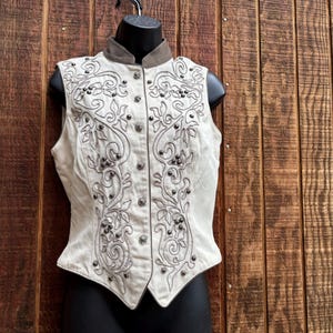 Puede incluir: Chaleco color crema con intrincados bordados florales y volutas en gris. El chaleco presenta un cuello alto, una parte delantera abotonada y tachuelas decorativas. El chaleco se exhibe en un maniqu&iacute; negro sobre un fondo de madera.