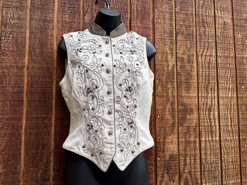Puede incluir: Chaleco color crema con intrincados bordados florales y volutas en gris. El chaleco presenta un cuello alto, una parte delantera abotonada y tachuelas decorativas. El chaleco se exhibe en un maniqu&iacute; negro sobre un fondo de madera.