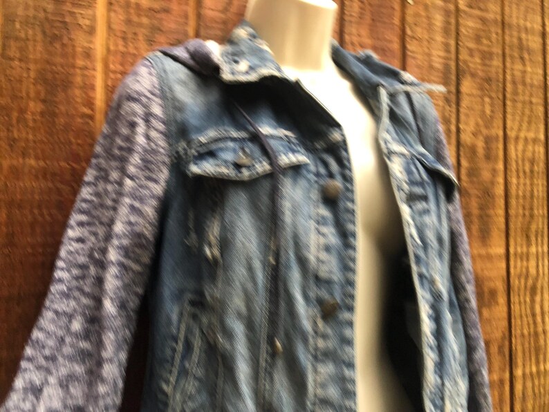 May include: A blue denim jacket with a hood and patterned sleeves. The jacket has a button-up front, two chest pockets, and a classic collar. The sleeves are a patterned knit fabric.