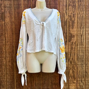 May include: White long-sleeved blouse with floral embroidery in yellow, blue, and green on the sleeves. The blouse has a tie-front closure and a V-neckline with blue stitching. The sleeves are long and gathered at the wrists.