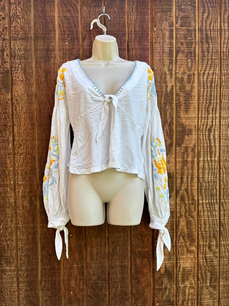 May include: White long-sleeved blouse with floral embroidery in yellow, blue, and green on the sleeves. The blouse has a tie-front closure and a V-neckline with blue stitching. The sleeves are long and gathered at the wrists.