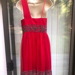 K&ouml;nnte beinhalten: Ein leuchtend rotes Kleid mit One-Shoulder-Design und zarten Spaghettitr&auml;gern. Das Kleid hat einen verzierten Bund und Saum mit aufwendiger Perlenarbeit. Der flie&szlig;ende, leichte Stoff erzeugt eine elegante Silhouette, perfekt f&uuml;r besondere Anl&auml;sse.