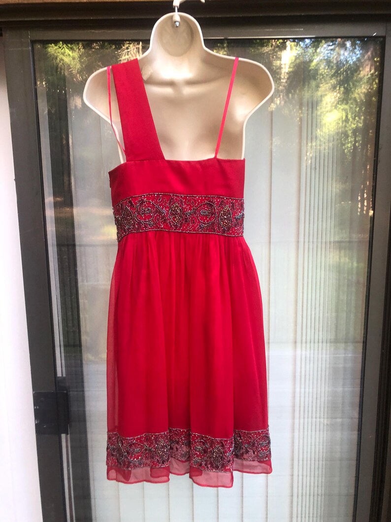 K&ouml;nnte beinhalten: Ein leuchtend rotes Kleid mit One-Shoulder-Design und zarten Spaghettitr&auml;gern. Das Kleid hat einen verzierten Bund und Saum mit aufwendiger Perlenarbeit. Der flie&szlig;ende, leichte Stoff erzeugt eine elegante Silhouette, perfekt f&uuml;r besondere Anl&auml;sse.