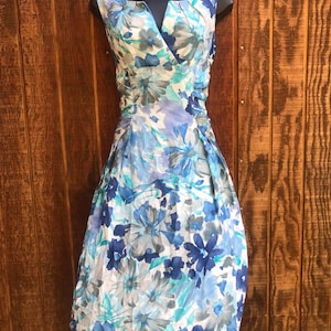 May include: A sleeveless floral dress in shades of blue, white, and gray. The dress features a V-neckline, gathered detailing at the waist, and a knee-length skirt. The floral pattern includes large, stylized blooms.
