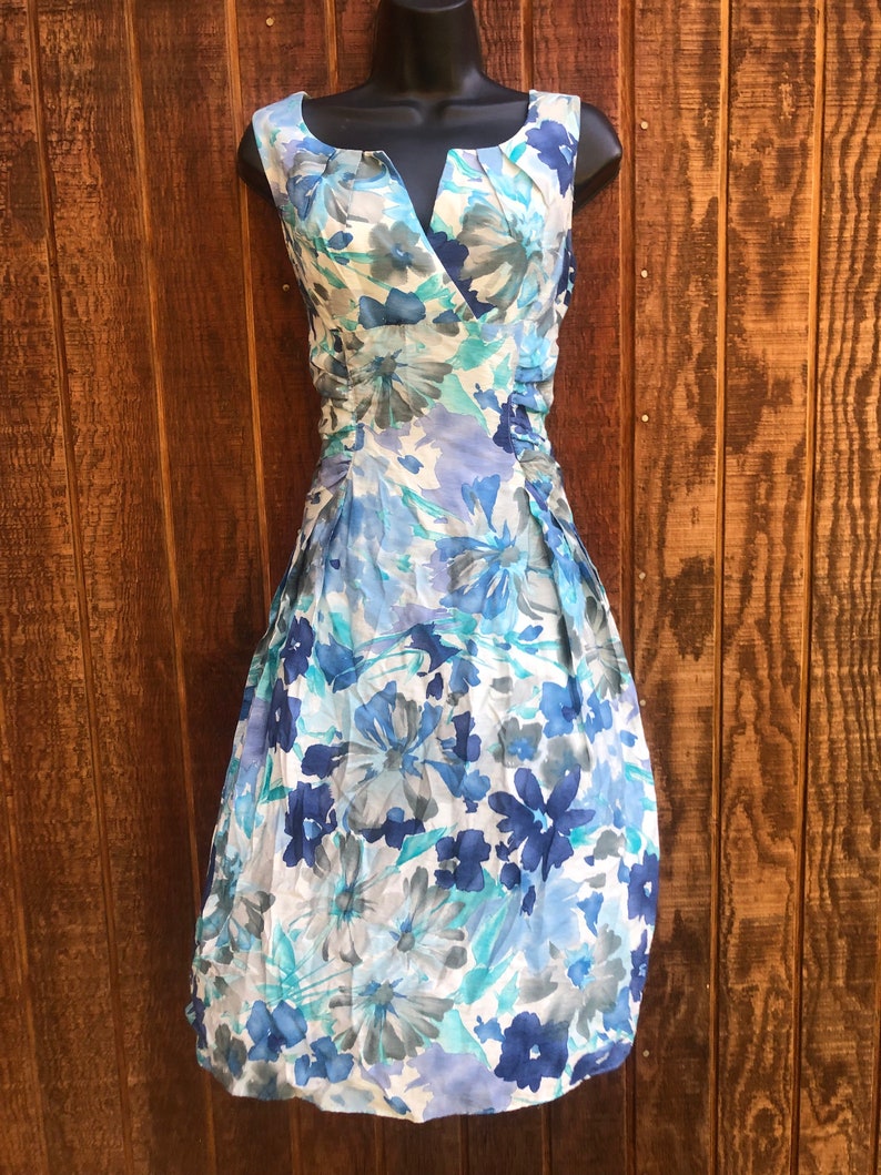 May include: A sleeveless floral dress in shades of blue, white, and gray. The dress features a V-neckline, gathered detailing at the waist, and a knee-length skirt. The floral pattern includes large, stylized blooms.