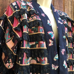 May include: A patchwork kimono-style robe with a dark blue butterfly print on the lapel and sleeves. The robe features layers of black fabric with a ruffled edge, and panels of floral and geometric patterns in pink, teal, and gold.