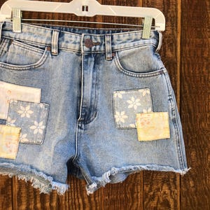 May include: Light blue denim shorts with frayed edges. The shorts feature three decorative patches: two with floral patterns and one with a vintage-style design. The shorts are hanging on a white hanger.