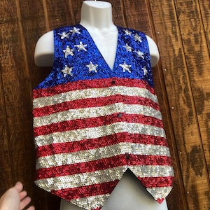 May include: A sequined vest in the style of the American flag. The top portion is blue with silver stars, and the lower portion features red and silver horizontal stripes. The vest has a V-neck and is buttoned down the front.