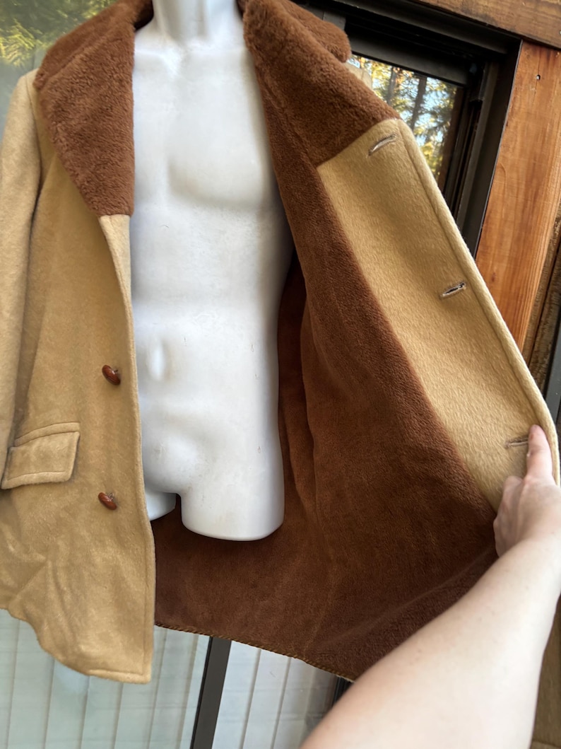Może przedstawiać: Beżowo-brązowy płaszcz w stylu vintage z kołnierzem i podszewką z imitacji futra. Płaszcz ma dwurzędowy kr&oacute;j z dwoma guzikami i kieszenią z przodu. Płaszcz jest otwarty, odsłaniając brązowe wnętrze.