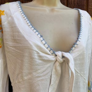May include: White long-sleeve top with a knotted front and a V-neckline. The neckline and sleeves are adorned with blue stitching. Floral embroidery in yellow and blue accents the sleeves. The top is displayed on a mannequin.