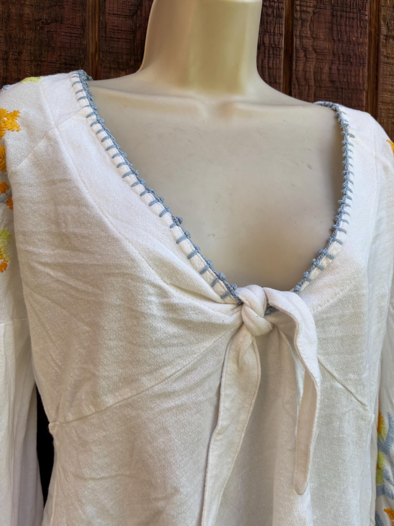 May include: White long-sleeve top with a knotted front and a V-neckline. The neckline and sleeves are adorned with blue stitching. Floral embroidery in yellow and blue accents the sleeves. The top is displayed on a mannequin.