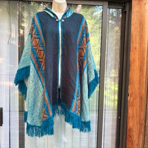 May include: A hooded poncho in shades of teal, navy, and orange, with a geometric pattern on the sleeves and a fringe hem. The poncho has a central zipper and wooden toggle closures. The garment is displayed on a mannequin.