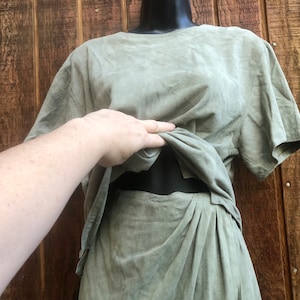 May include: A two-piece sage green outfit on a mannequin. The top is a short-sleeved, boxy style, and the skirt is a wrap style with pleats. The outfit is made of a soft, textured fabric. The background is a wooden wall.