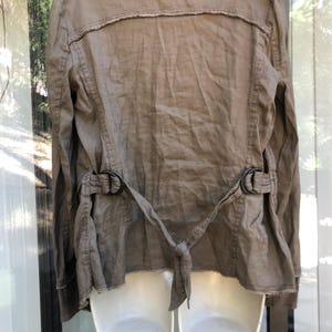 K&ouml;nnte beinhalten: Eine beige Leinenjacke mit langen &Auml;rmeln und einem Reverskragen. Die Jacke hat einen G&uuml;rtel mit zwei schwarzen Metallschnallen und einen Bindverschluss in der Taille. Die R&uuml;ckseite der Jacke hat ein ausgefranstes Detail.