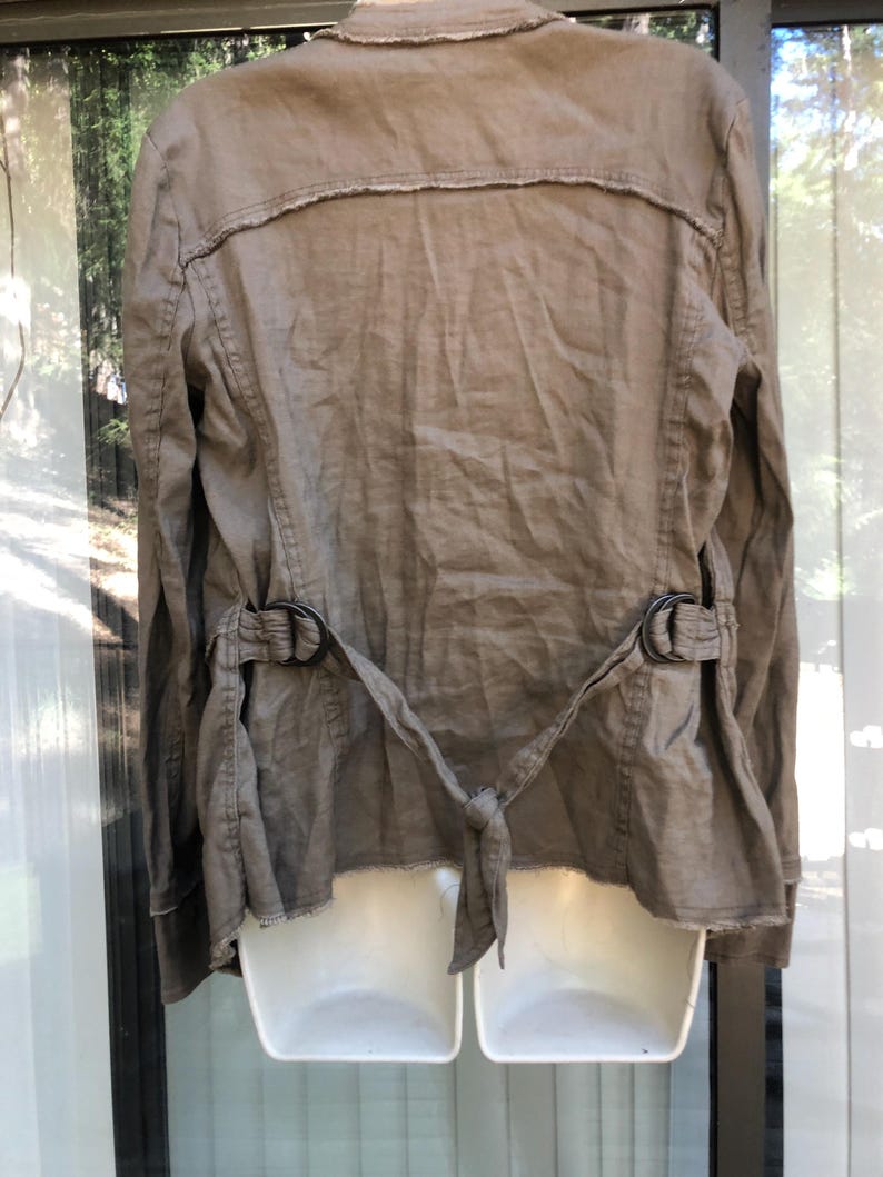 K&ouml;nnte beinhalten: Eine beige Leinenjacke mit langen &Auml;rmeln und einem Reverskragen. Die Jacke hat einen G&uuml;rtel mit zwei schwarzen Metallschnallen und einen Bindverschluss in der Taille. Die R&uuml;ckseite der Jacke hat ein ausgefranstes Detail.