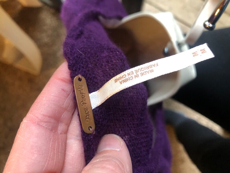 May include: A close-up of a purple garment with a tag that says "free people". A white tag with the text "MADE IN CHINA" and "FABRIQUE EN CHINE" is also visible. The garment is held by a person.