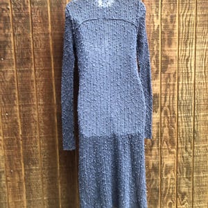K&ouml;nnte beinhalten: Ein langes, blaues, strukturiertes Kleid mit langen &Auml;rmeln h&auml;ngt vor einem h&ouml;lzernen Hintergrund. Das Kleid hat ein einzigartiges, erhabenes Punktmuster im gesamten Stoff. Das Kleid h&auml;ngt an einem wei&szlig;en Kleiderb&uuml;gel.