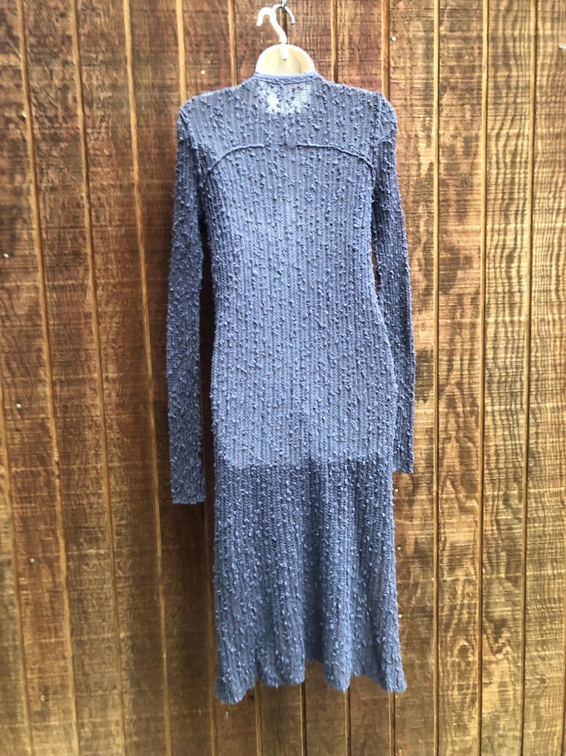 K&ouml;nnte beinhalten: Ein langes, blaues, strukturiertes Kleid mit langen &Auml;rmeln h&auml;ngt vor einem h&ouml;lzernen Hintergrund. Das Kleid hat ein einzigartiges, erhabenes Punktmuster im gesamten Stoff. Das Kleid h&auml;ngt an einem wei&szlig;en Kleiderb&uuml;gel.