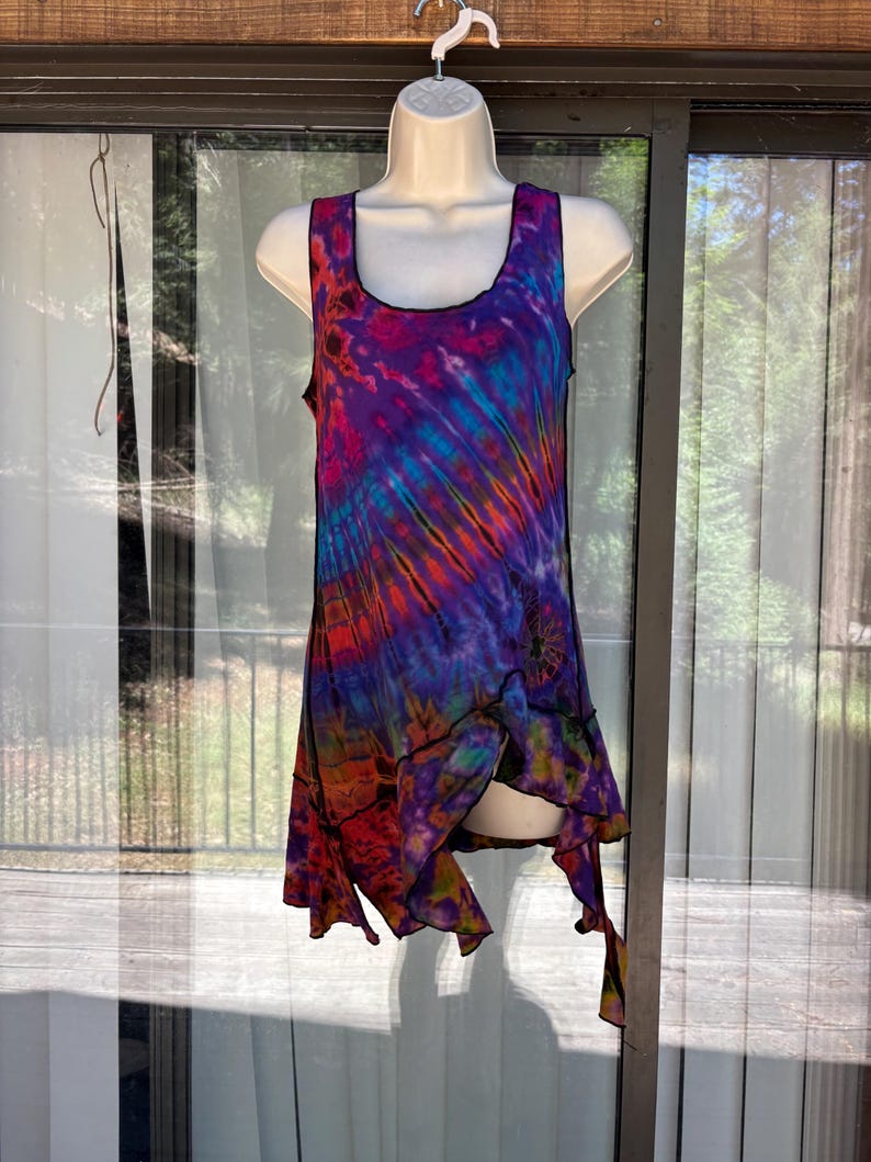 May include: A sleeveless tunic top featuring a vibrant tie-dye pattern in shades of purple, blue, orange, and pink. The garment has a scoop neckline, black stitched seams, and an asymmetrical hemline with multiple points that drape unevenly.
