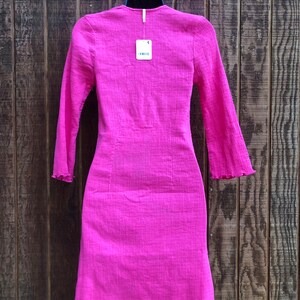 May include: A bright pink, long-sleeved dress with a textured fabric and a ruffled hem. The dress is hanging on a black hanger against a wooden background. A price tag is attached to the dress.