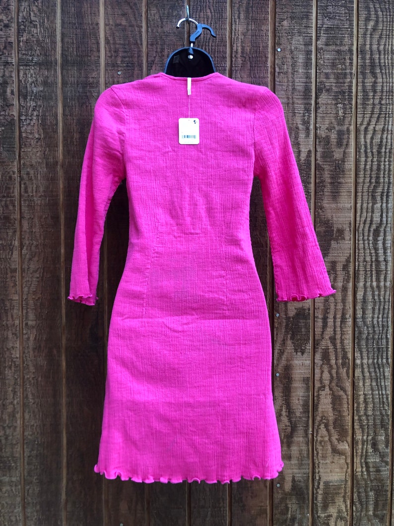 May include: A bright pink, long-sleeved dress with a textured fabric and a ruffled hem. The dress is hanging on a black hanger against a wooden background. A price tag is attached to the dress.